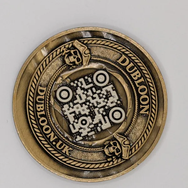 The face of a struck antique-brass Dubloon: a QR mark with bone-white modules, DUBLOON above and DUBLOON.UK below, two small skulls, a rope edge
